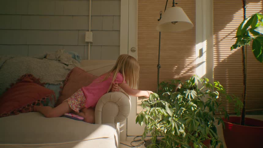 A blonde girl lies on a couch in a sunlit room, curiously touching the leaves of a nearby potted plant