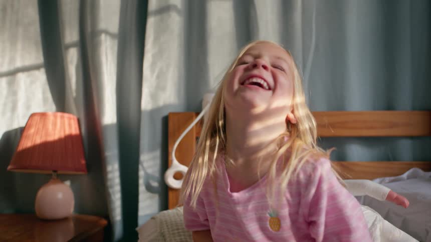 A cute little blonde girl in pink pajamas sits on her bed in the morning sunlight and makes silly faces at the camera