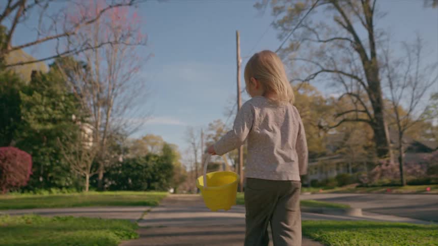 A cute little toddler carries a yellow bucket while walking down a suburban sidewalk on a beautiful, sunny spring day