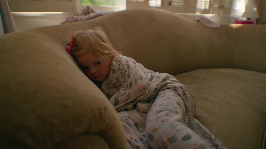 An unwell-looking little blonde girl in her pajamas lies on a large plush couch wrapped in a blanket, staring sadly at the camera