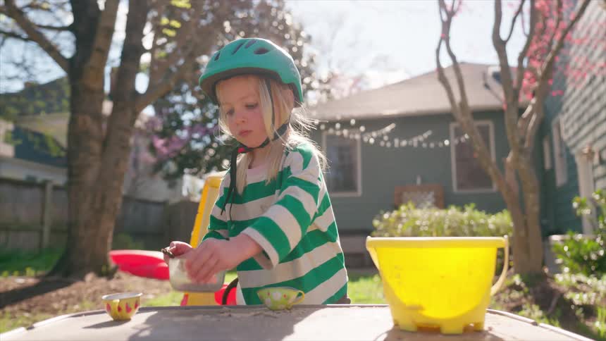 A young child wearing a bicycle helmet carefully scoops sand into a toy teacup while playing in their backyard on a sunny day