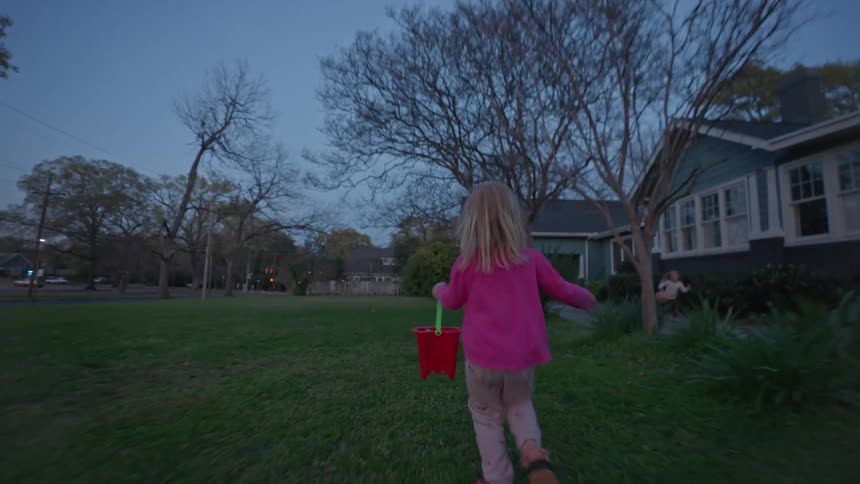 Two young sisters joyfully run with their baskets across the front lawn of their home at twilight