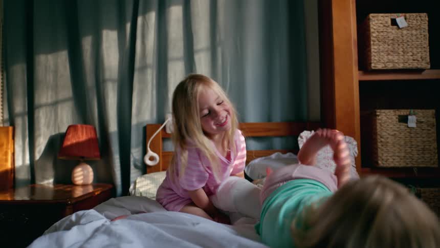 Two adorable sisters in pajamas laugh and play on a bed in their sunlit bedroom in slow motion
