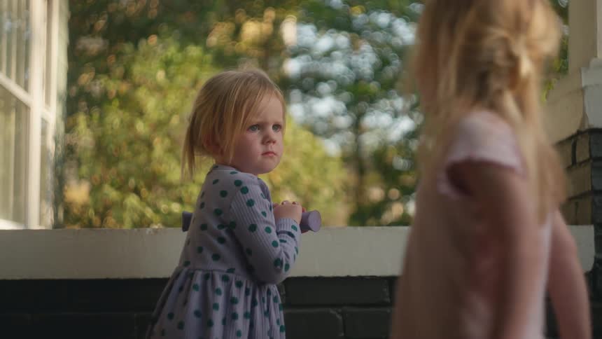A slow focus pull reveals a young girl with a solemn expression watching her sister play on a sunny porch