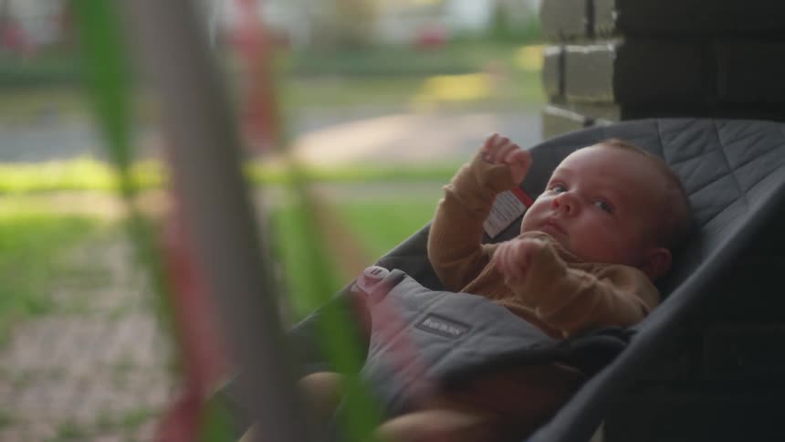 A young girl with blonde hair gently interacts with her newborn sibling relaxing in a bouncer on the front porch