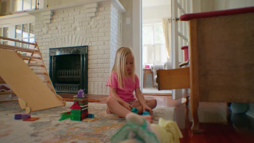 A young blonde girl in a pink shirt plays with colorful building blocks on a rug in her sunlit living room