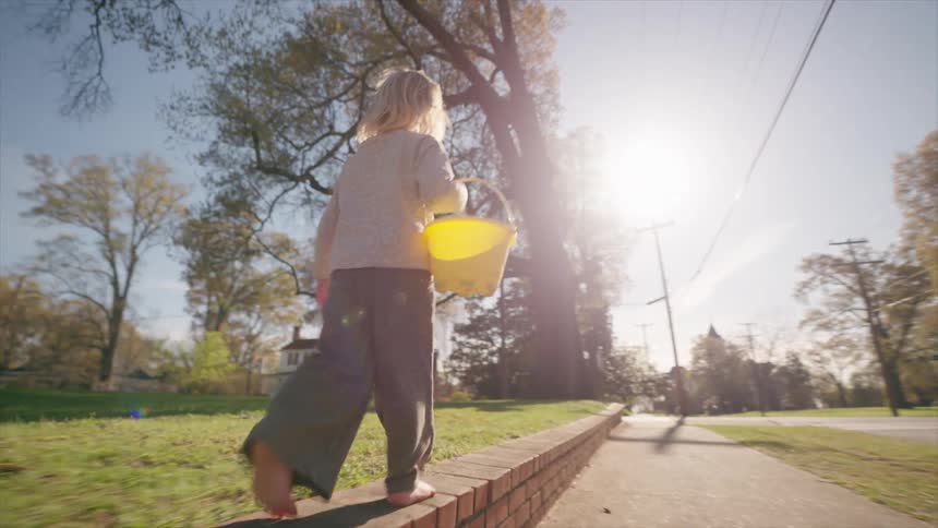 A barefoot child with a yellow bucket balances and walks along a low brick wall in a sunlit neighborhood