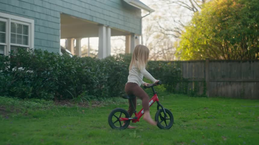 A young girl practices riding her red balance bike in the backyard, takes a tumble onto the grass, smiles, and determinedly gets back up to ride again