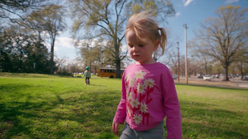A pensive little girl in a pink floral shirt walks slowly through her front yard on a sunny day
