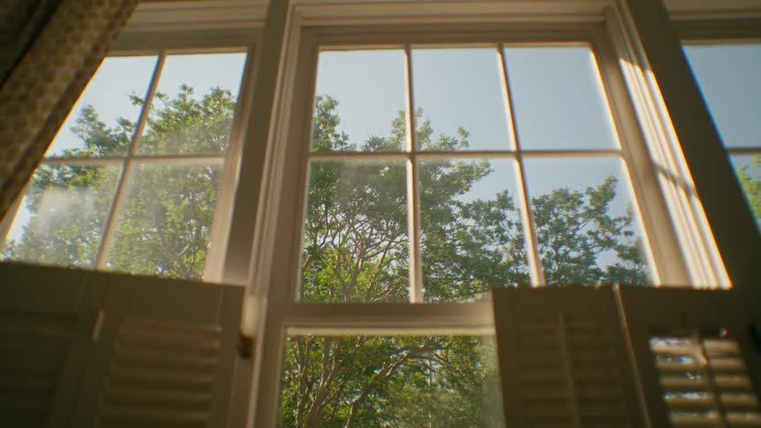 Sunlight shines through an open window with shutters, revealing green trees outside on a beautiful day