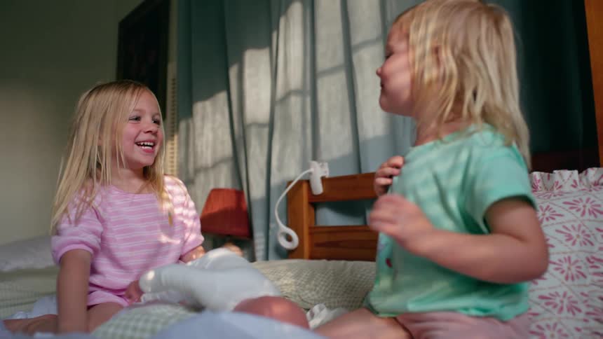 Adorable blonde sisters laugh and play together in their sun-drenched bedroom