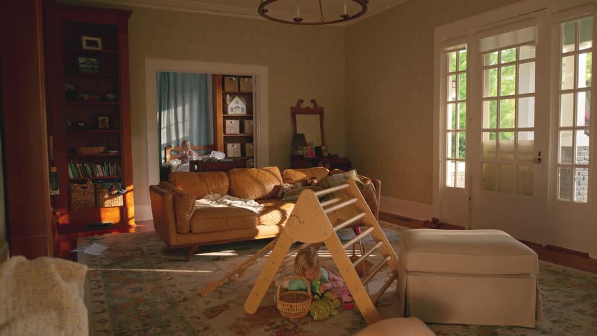 A toddler plays quietly in a sunlit and messy family living room while their sibling is seen in the adjoining room