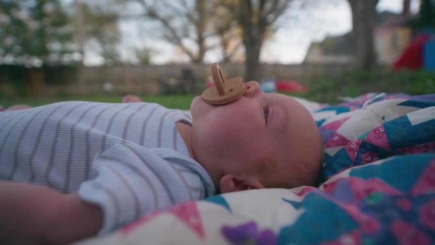 A cute newborn baby with blue eyes and a pacifier lies peacefully on a patterned quilt in a backyard