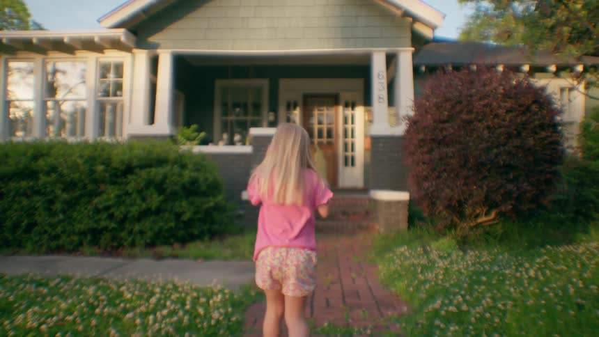 Little girl in pink walks up the brick path toward a suburban family home at golden hour