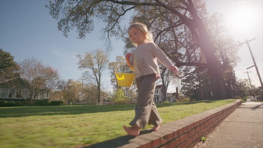 A barefoot toddler with a yellow bucket carefully balances while walking on a brick wall, then jumps off and runs away on a sunny day
