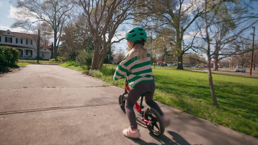 A little girl wearing a helmet rides her balance bike down a neighborhood sidewalk on a sunny day