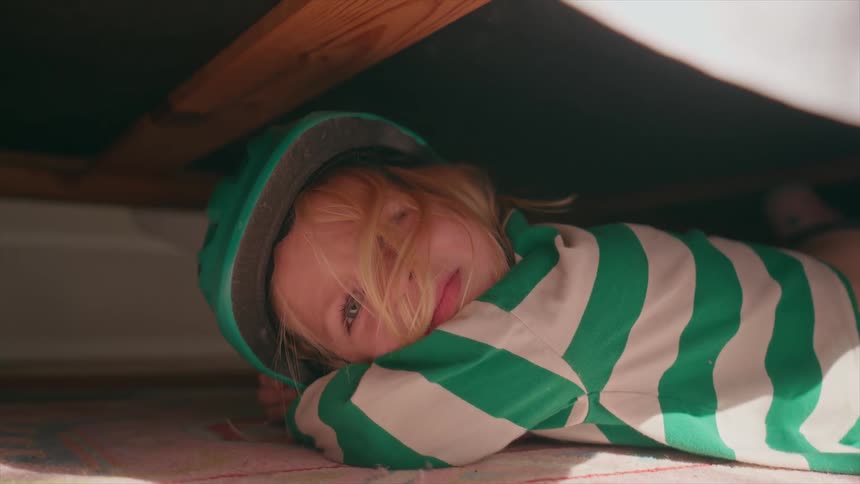 Young child in a green helmet lies under a bed indoors with a playful, curious expression