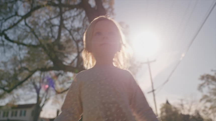 A cinematic portrait of a young blonde girl standing in a sun-drenched neighborhood as the camera looks up at her