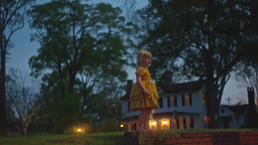 A barefoot little girl in a yellow princess dress stands and walks on a low brick wall in her front yard at twilight
