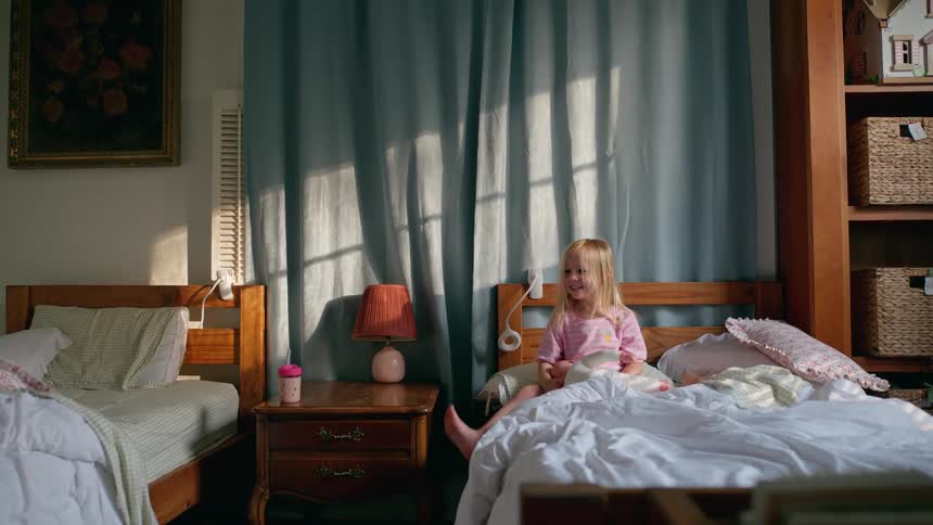 A happy little girl in pink pajamas plays with her stuffed animal in her sunlit bedroom in the morning