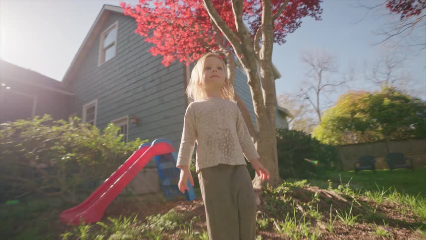 A curious toddler girl walks barefoot through her sunny, green backyard in a beautiful slow-motion tracking shot