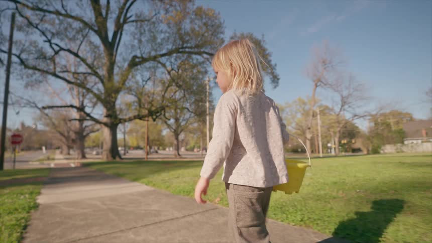 A young barefoot child with a yellow bucket embarks on a sunny walk down a quiet suburban street