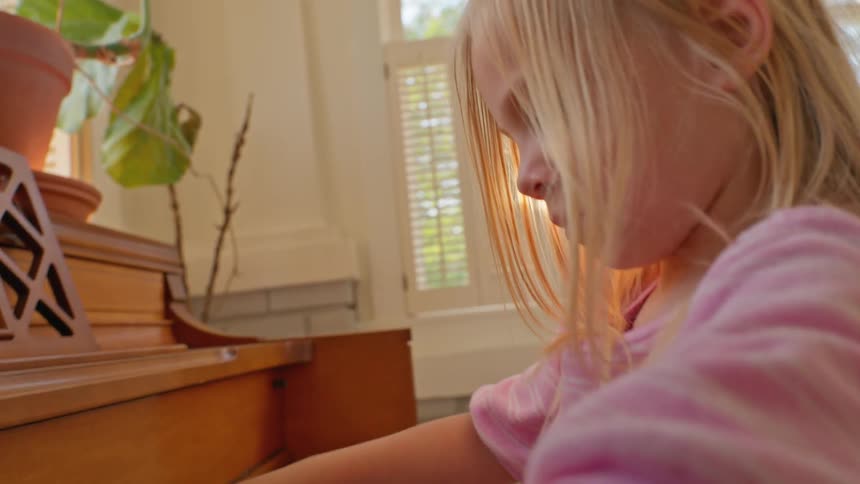 A young blonde girl bathed in morning sunlight intently plays a vintage wooden piano in her home