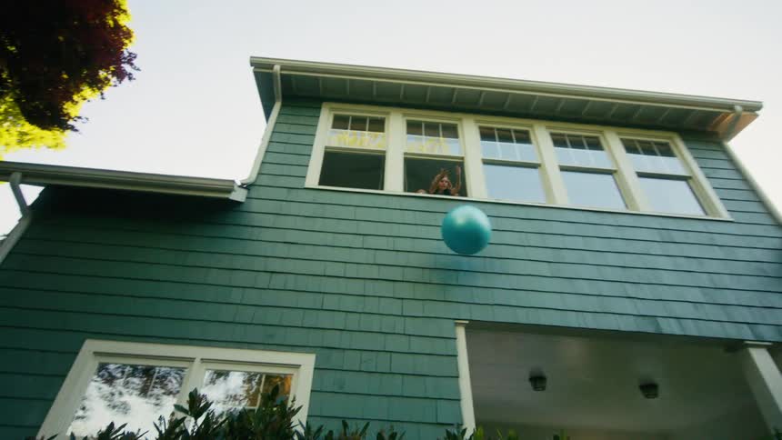 A young boy throws a ball up to a girl in a second-story window of their house