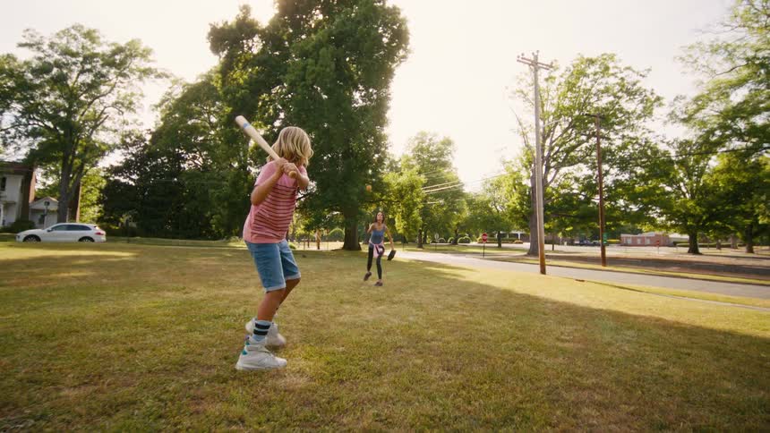 A mother pitches a baseball to her young son, who hits the ball while playing in a sunny park