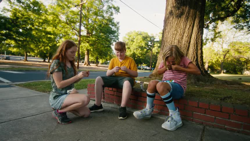 Three kids sit together on a sidewalk under a tree, carefully unwrapping small items on a sunny afternoon