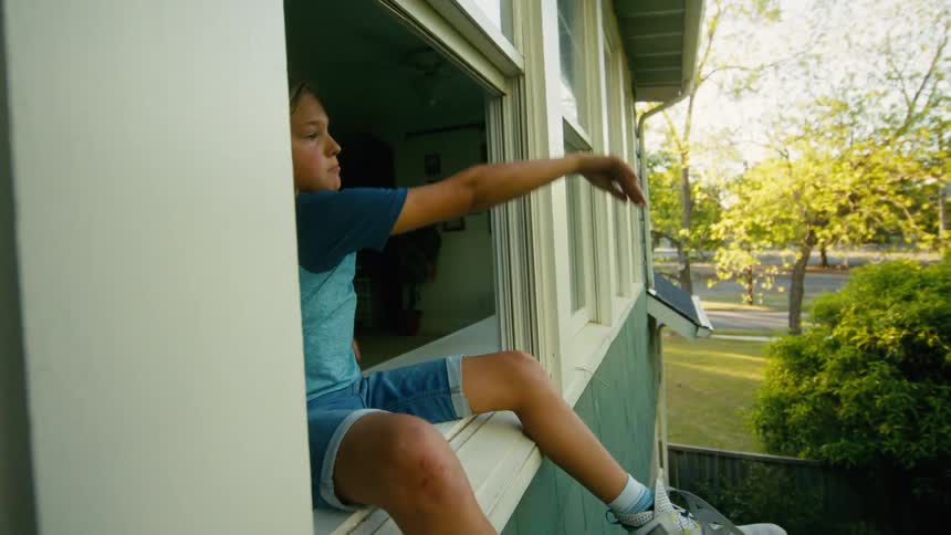 A young boy sitting on a windowsill throws a large blue exercise ball out of the window