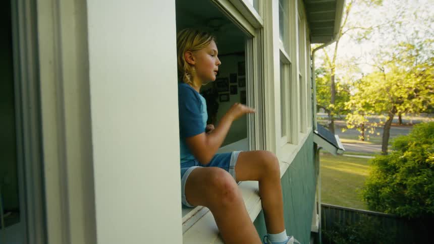 A young boy with a scraped knee sits in an open window on a summer day and throws a ball out to play