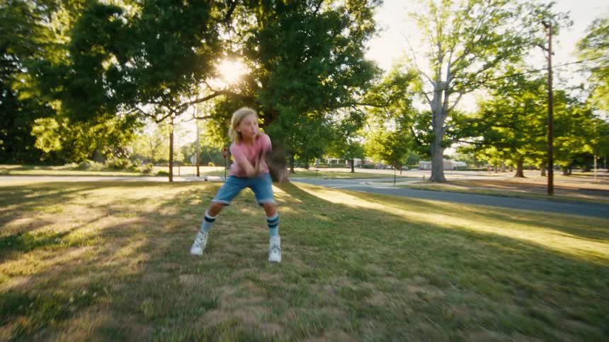 A young boy with long blonde hair joyfully catches and throws a football in a sunlit park during a beautiful summer day