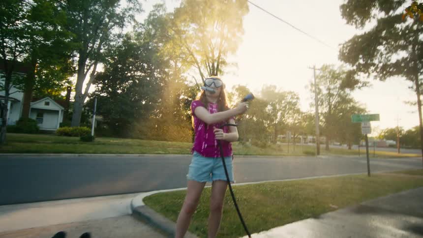 A young girl in a snorkel mask playfully sprays her friends with a hose as they pretend to swim on a sunny driveway
