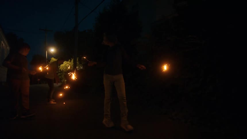 Three kids walk down a dark street at night, waving glowing sparklers