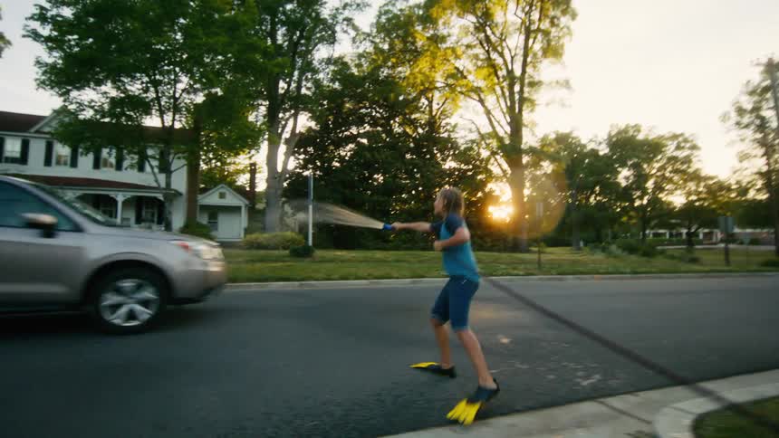 Playful kids wearing flippers have a summer water fight and spray a passing car with a garden hose at sunset