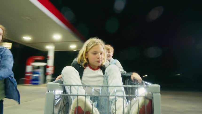 A point-of-view shot from a shopping cart as a group of excited kids race through a gas station parking lot at night