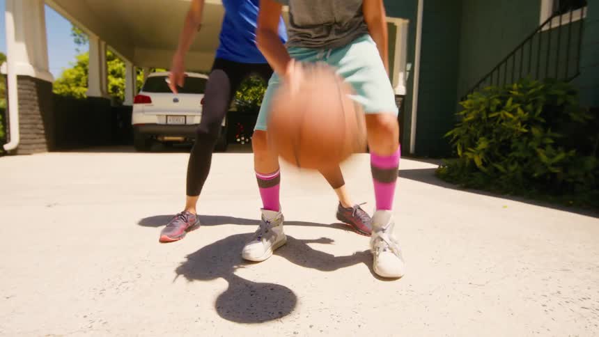 A young boy dribbles a basketball while his mother plays defense in the driveway, ending with a shot at the hoop
