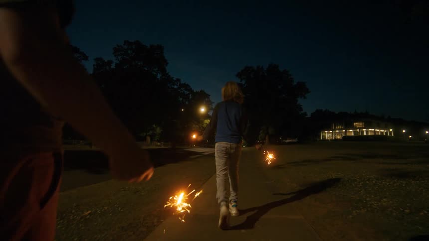 Following a group of kids as they run down a sidewalk at night, joyfully waving lit sparklers