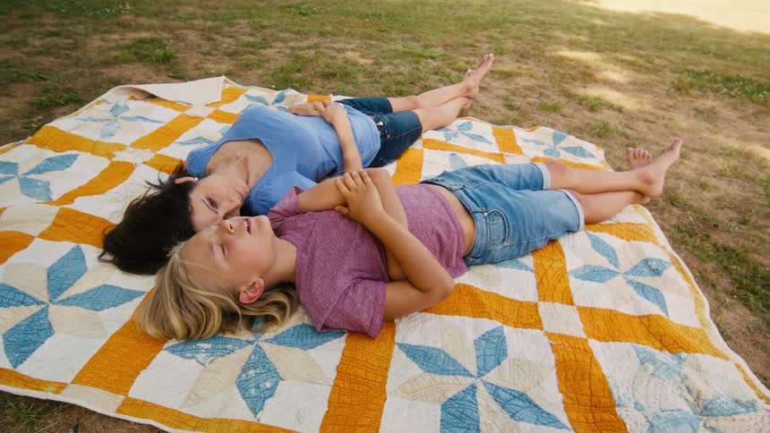 A mother and her young son share a quiet, peaceful moment talking and relaxing while lying on a colorful quilt in a sunlit park