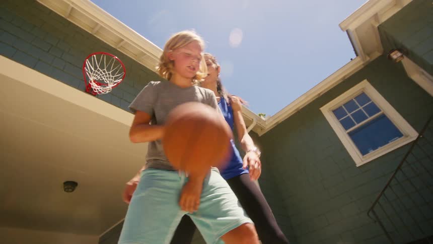 Low-angle slow motion shot of a boy dribbling past his mom to make a successful basketball shot in their backyard on a sunny day