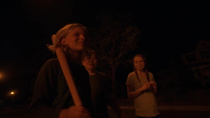 Three young friends on a suburban street at night, with one boy smiling and swinging a baseball bat from a low angle