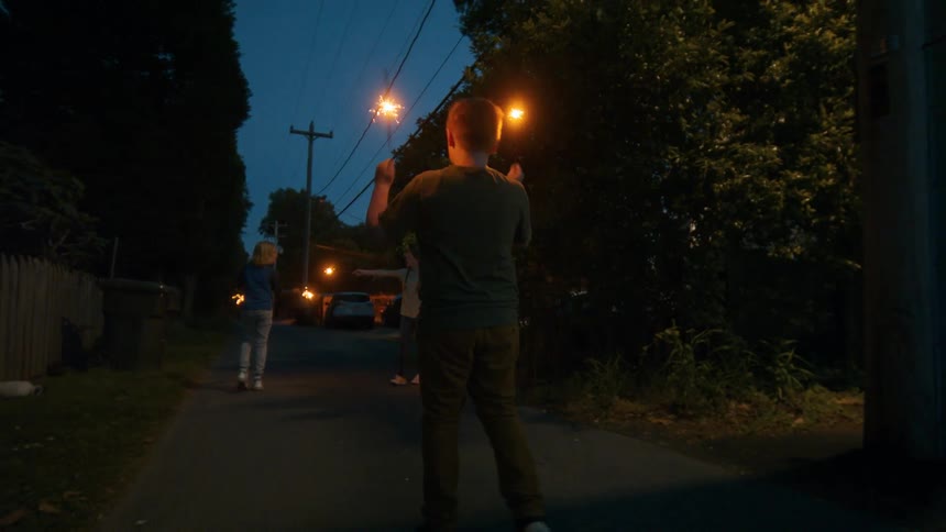 Three children walk down a suburban street at dusk, waving lit sparklers in celebration