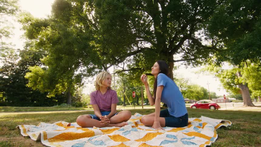 A happy mother and her son relax on a colorful quilt in a sunlit park, eating apples and enjoying a conversation
