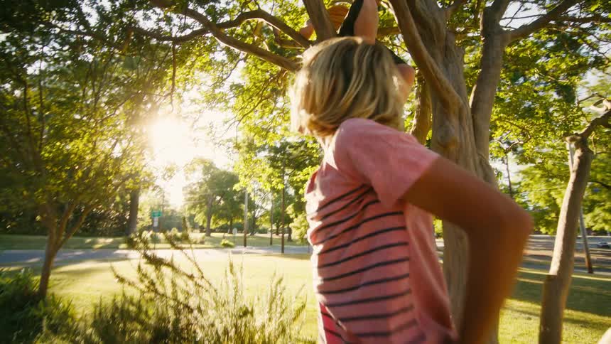 A boy with long blonde hair jumps down from a tree to meet his friends in a sunny park