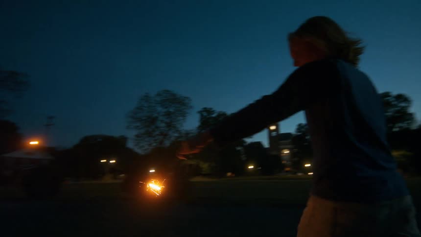 A tracking shot follows three joyful children running and dancing in a park at twilight while playing with bright sparklers