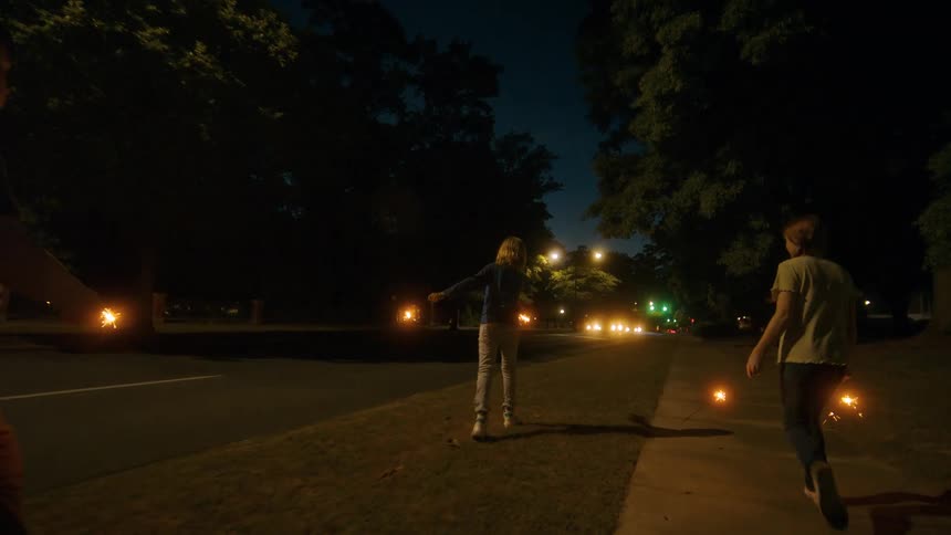 A group of kids celebrate by walking down the sidewalk at night, lighting up the darkness with sparklers