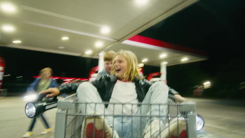 POV of an adventurous boy holding two bright flashlights while riding in a shopping cart pushed by his friends at night
