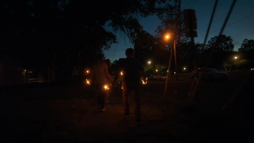 A group of children run through a neighborhood street at dusk holding sparklers
