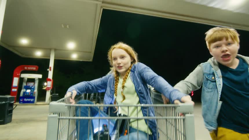 Joyful kids have fun pushing a friend in a shopping cart through a gas station at night